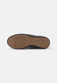 Suela de zapatilla negra con una base de goma marrón, que presenta un patrón de agarre texturizado y un logo en relieve. Bordes suaves y forma redondeada.