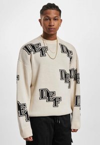 Cremfarbener Strickpullover mit schwarzen "DEF" Logos in verschiedenen Größen, oversized Passform, gerippte Manschetten und Rundhalsausschnitt. Kombiniert mit schwarzen Hosen.