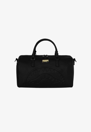 Sac de sport cylindrique noir avec motif en relief de bouche de requin et deux poignées courtes, avec une petite étiquette métallique dorée près de la fermeture éclair.