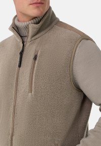 Mees, kes kannab beeži sherpa fliisist vestikut, mille all on heledast beežist kõrge kaelusega sviiter, lukustatava rinna taskuga ning nahast õlaplekkidega.