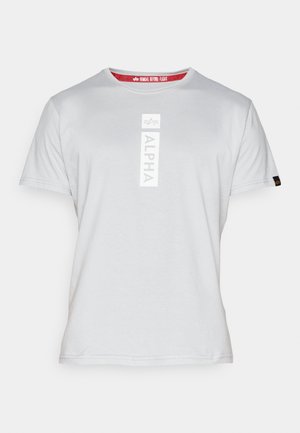 T-shirt en coton gris avec un logo vertical "ALPHA" en blanc sur le devant, un col rond et une étiquette rouge à l'intérieur avec "ENLEVER AVANT LE VOL."