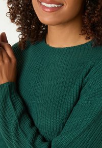 Grüner Strickpullover mit geripptem Muster, Rundhalsausschnitt und langen Ärmeln mit lockeren Bündchen, vor einem neutralen Hintergrund präsentiert.
