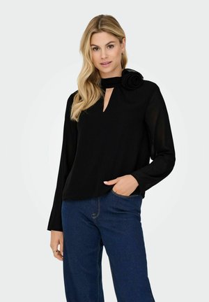 Femme aux longs cheveux blonds portant un haut noir à manches longues avec un détail de rose au niveau du col et un jean bleu, posant avec une main dans la poche.