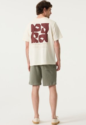 T-shirt met korte mouwen in crèmekleur, met een bordeauxrode afbeelding op de achterkant, gecombineerd met groene korte broek en beige instapschoenen.