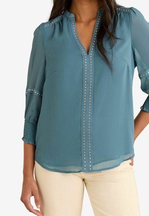 Femme portant un chemisier bleu sarcelle avec des clous argentés le long de l'encolure, des épaules et des manches, associé à un pantalon beige clair.