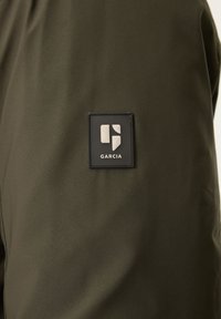 Olijfgroen stof met een gladde textuur, voorzien van een rechthoekig zwart patch met een wit logo met de tekst "GARCIA" aan de linkerkant.