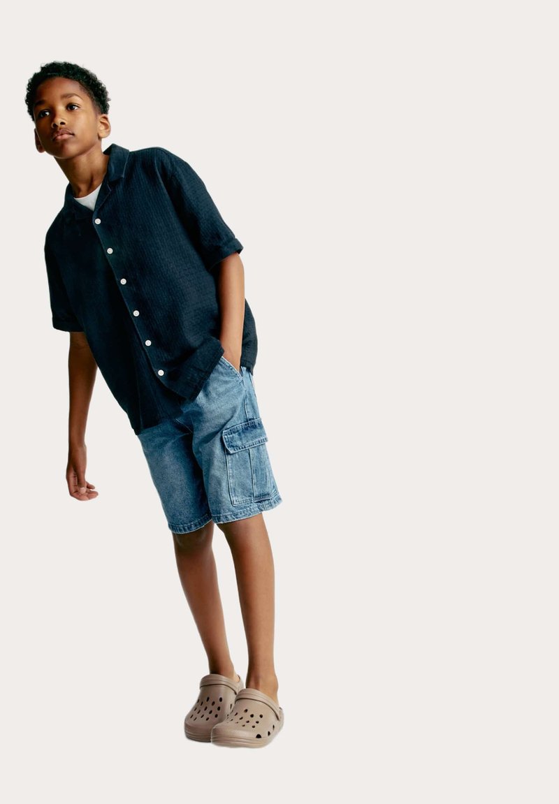Una camicia nera a maniche corte con bottoni, pantaloni cargo in denim e zoccoli beige con fori di ventilazione indossati da un ragazzo che si appoggia leggermente.