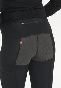 Mustat legginsit, joissa on teksturoidusta harmaasta paneelista ja pieni punainen emblem. Takana on vetoketjullinen tasku ja tyylikäs muotoilu.