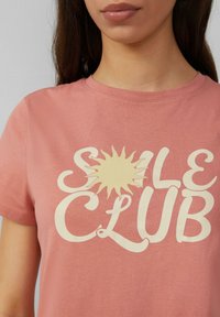 Korte mouwen, blushroze katoenen t-shirt met een grote, crèmekleurige grafische opdruk die "SOLE CLUB" leest met een zonillustratie.