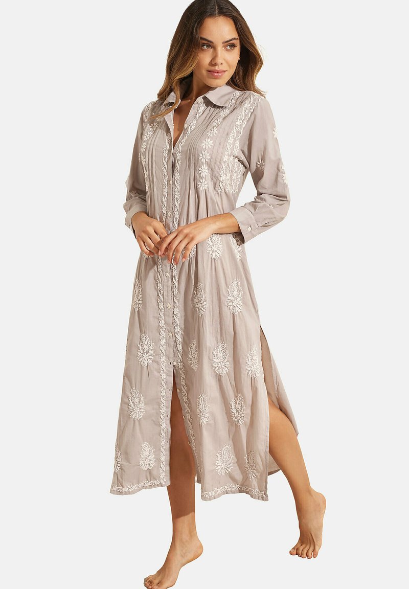 Robe longue à boutons en tissu beige clair ornée de broderies florales blanches et de fentes latérales. Avec un col, des manches longues et une coupe décontractée.
