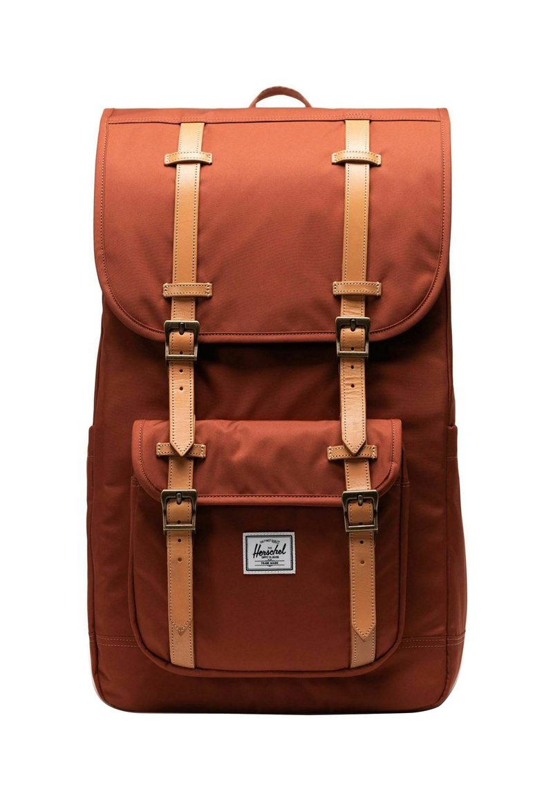 Sac à dos en tissu marron avec des sangles et des boucles en cuir, poche avant et rabat supérieur. Présente une étiquette de marque à l'avant.