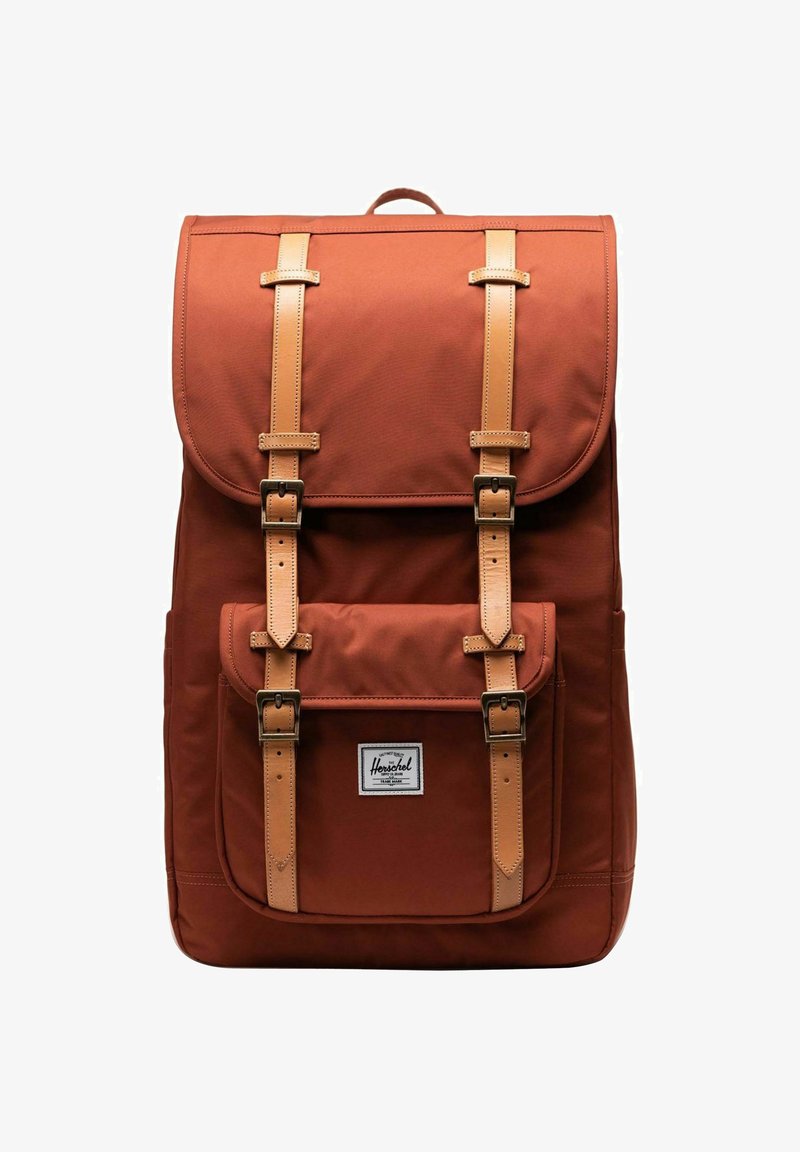 Sac à dos en tissu marron avec des sangles et des boucles en cuir, poche avant et rabat supérieur. Présente une étiquette de marque à l'avant.