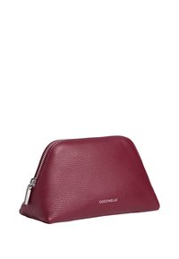 Czerwona skórzana kosmetyczka o kształcie trójkąta, teksturowana powierzchnia, metalowy zamek błyskawiczny oraz wytłoczone logo marki z przodu.