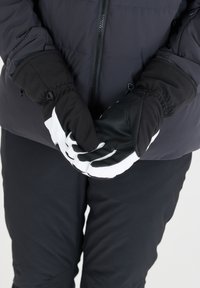 Svartvita skidhandskar med texturerat grepp, kontrasterande tumme och åtsittande handledsmuddar, i kombination med en mörk isolerad jacka.