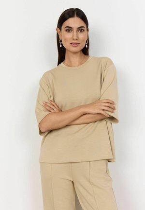 Camiseta oversized color beige claro, fabricada en tejido suave, con cuello redondo, mangas cortas y un branding sutil. Combinada con pantalones a juego.