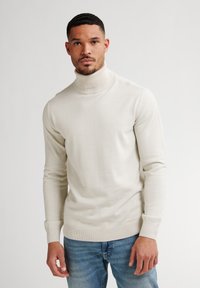 Jersey de cuello alto crema con mangas largas, puños y dobladillo acanalados, combinado con jeans azules, con una textura ajustada y suave.