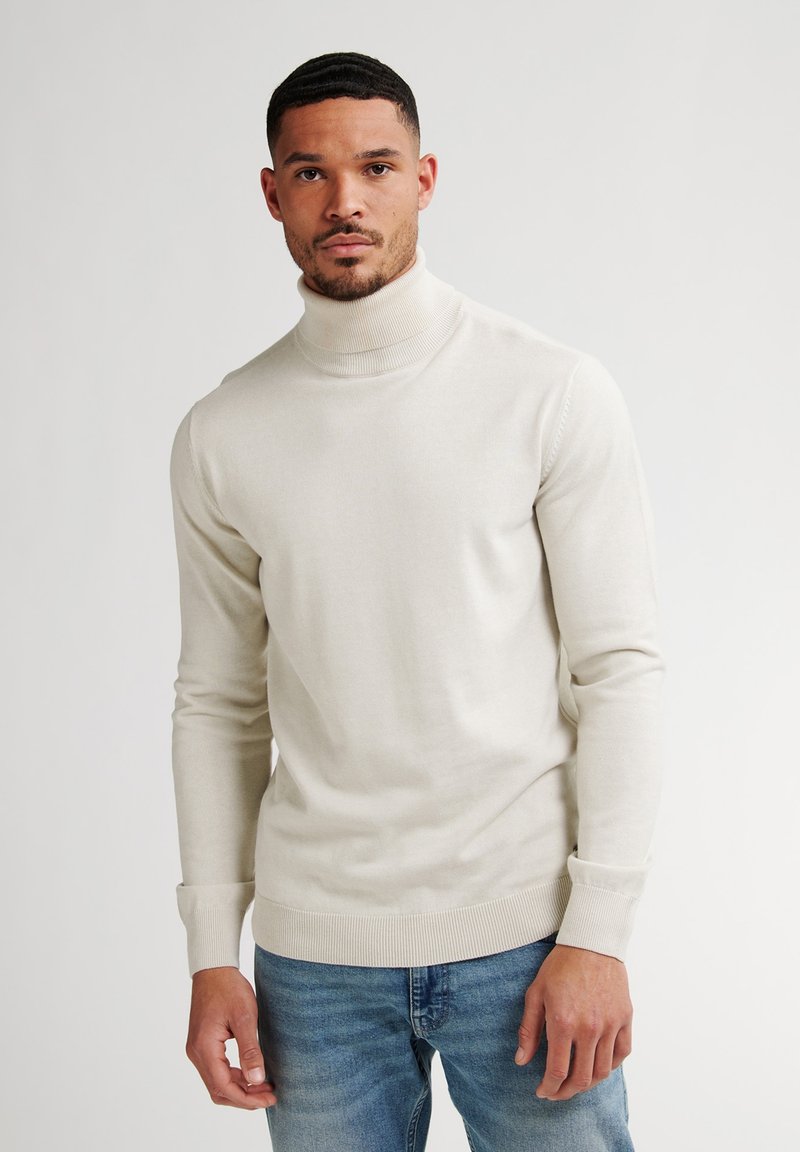 Jersey de cuello alto crema con mangas largas, puños y dobladillo acanalados, combinado con jeans azules, con una textura ajustada y suave.