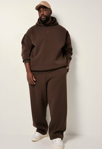Bruine hoodie met een losse pasvorm, gecombineerd met bijpassende bruine joggingbroek. Accentdetails omvatten een logo en een lichtgekleurde pet.