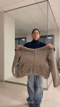 Γυναίκα με μαύρα μαλλιά που φοράει κρίκους στα αυτιά και σκούφο πουλόβερ κρατά ένα μπεζ φλις μπουφάν με μπροστινές τσέπες μέσα σε ένα δωμάτιο με καθρεφτισμένους τοίχους.