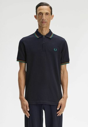 Uomo con capelli corti e scuri indossa una polo blu navy con colletto e maniche a righe verdi e arancioni, in piedi davanti a uno sfondo bianco uniforme.