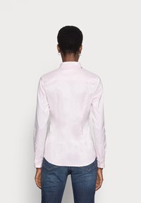 Persona con capelli corti e ricci, che indossa una maglietta a maniche lunghe aderente di un rosa chiaro e jeans blu, in piedi di spalle su uno sfondo neutro.