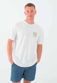 Camiseta blanca de manga corta con un logo circular y un diseño de sol en el pecho, combinada con pantalones cortos azules. Fondo simple, corte relajado.