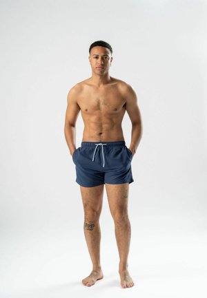 Homme torse nu portant un short de bain bleu marine avec poches, debout pieds nus sur un fond blanc, les mains dans les poches, face à l'avant.