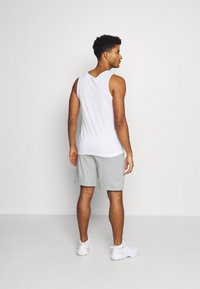 Vit ärmlös linne med rundad fåll, i kombination med grå shorts. Personen bär vita sneakers och står vänd bort mot en neutral bakgrund.