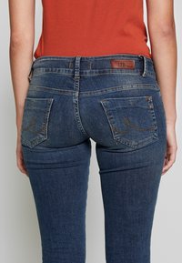 Jeans i denim med en kroppsnära passform, i mörkblått, med bakfickor med dekorativ sömmar och en brun läderlogga vid midjan.