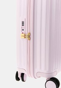 Valigia rigida rosa con righe verticali, zip dorate, un lucchetto a combinazione e quattro ruote nere per la mobilità.