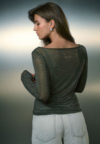 Maglia lunga a maniche lunghe di colore verde scuro trasparente adornata con piccoli borchie metalliche, caratterizzata da una scollatura ampia e un design aderente, abbinata a jeans grigio chiaro.