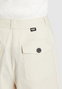 Beige linnen broek met een zijzak met een zwarte knop en een gestikt label met "khujo" branding op de tailleband.