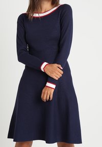 Navy dress with long sleeves, ribbed texture, and contrasting red and white stripes at the collar and cuffs. A-line silhouette.