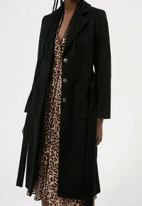 Manteau en laine noire au design cintré, avec des revers crantés, quatre boutons, poches avant et ceinture intégr�ée portée sur une robe à imprimé léopard.