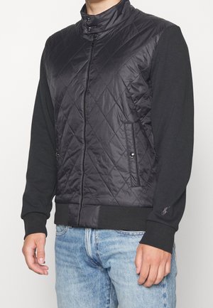 Homme portant une veste matelassée noire zippée avec poignets et col côtelés, associée à un jean bleu clair.