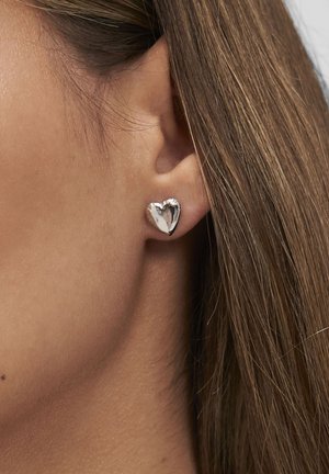 Boucle d'oreille stud en argent en forme de cœur portée sur l'oreille d'une femme avec des cheveux châtain clair visibles.