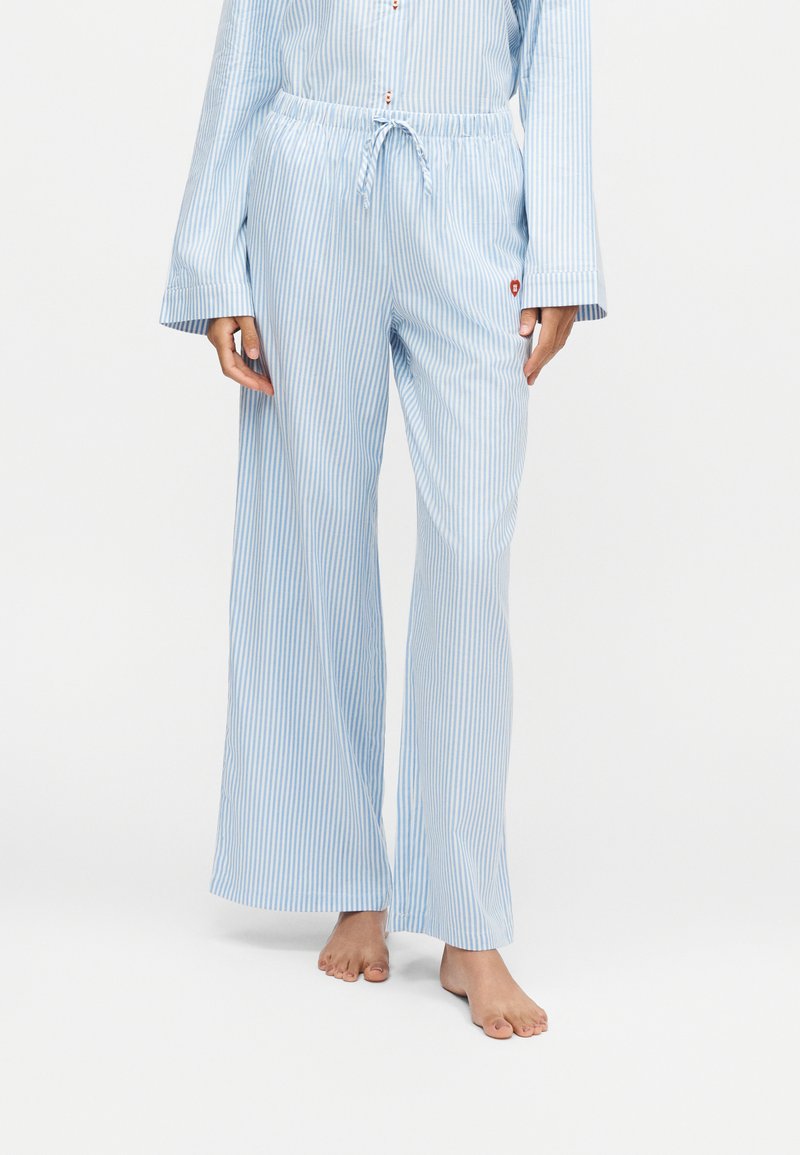 Personne portant un pyjama ample à rayures bleu clair et blanc, à manches longues et pantalon, debout pieds nus sur fond blanc.