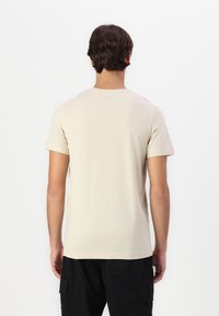 Gai bēšu īsām piedurknēm T-krekls ar apaļu izgriezumu, gluda tekstūra un vienkāršs dizains, redzams no aizmugures uz balta fona.