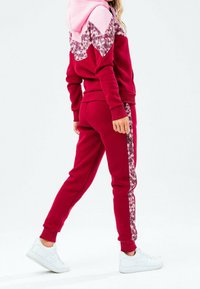 Chándal rojo con un diseño rosa y floral en la capucha. Presenta una silueta ajustada y acentos blancos en los pantalones. Zapatillas blancas.