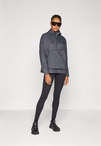 Lettet sort hættejakke med halvlynlås, kombineret med sorte tights og sorte træningssko. Glat tekstur, minimalistisk branding.