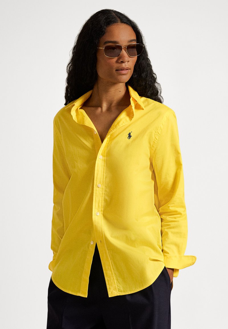 Moteris su ilgais garbanotais plaukais, dėvinti akinius nuo saulės, ryškiai geltoną marškinių tipo palaidinę su polo logotipu ir tamsias kelnes, stovinti su rankomis kišenėse.