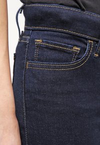 Bolsa y cinturilla de unos vaqueros de denim azul oscuro con costuras amarillas, usados por una persona con un brazo visible en el lado izquierdo.