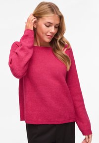 Pull en maille rose vif avec une texture côtelée, un col en forme de U et des manches longues. Il présente une coupe décontractée et des poignets côtelés.