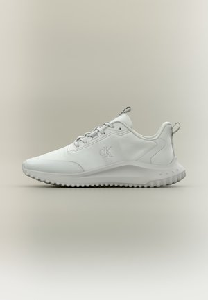 Baskets blanches basses avec semelle texturée, lacets à l’avant et logo « cK » sur le côté, présentées de profil sur un fond neutre.