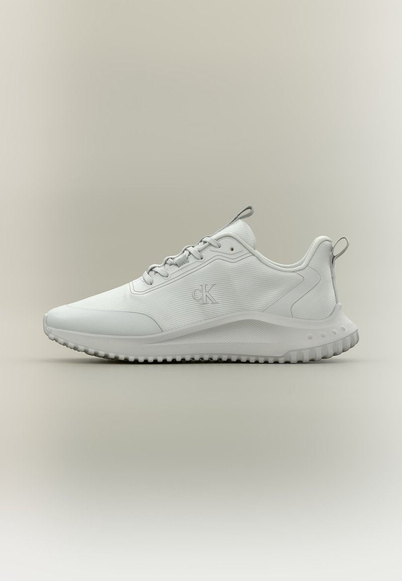 Fehér, alacsony szárú sportcipő texturált talppal, fűzővel az elején, és „cK” logóval az oldalon, oldalnézetben neutrális háttéren.