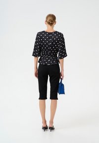 Crna bluzica s točkicama i peplum strukom, kombinirana s crnim capri hlačama. Model drži malu plavu torbicu i nosi crne sandale na petu.