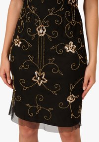 Černá sukně s zlatými flitrovými květinovými a vinnými výšivkami, průhledným lemem a rukama jemně položenými po stranách.