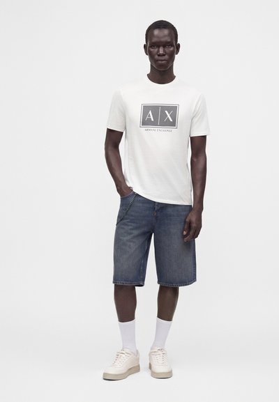 Мъж, облечен с бяла тениска Armani Exchange, сини дънкови къси панталони, бели чорапи и светли спортни обувки, стои пред едноцветен фон.