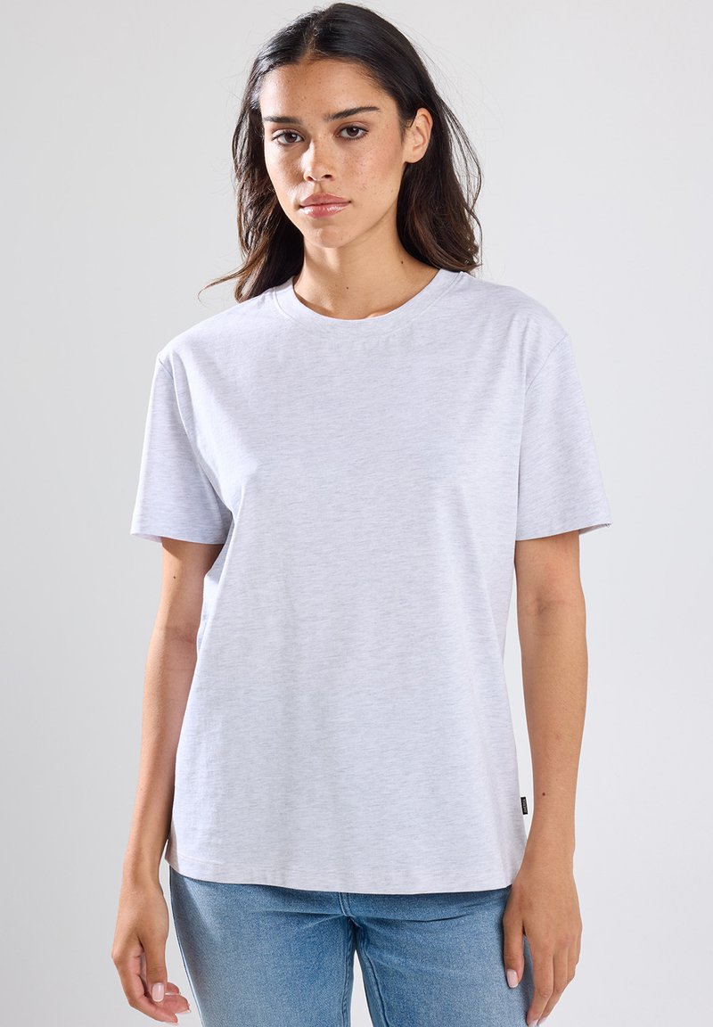 Femme aux longs cheveux bruns portant un t-shirt gris clair à manches courtes et un jean bleu, se tenant devant un fond gris clair.