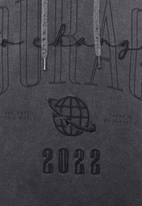 Sudadera gris con un gran diseño bordado en negro que dice "Courage" y un globo, con texto adicional abajo y cordones en la capucha.